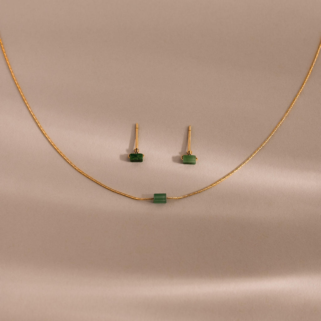 The Jade Bar Jewelry Set features a gold necklace and earrings with petite green jade stones, displayed on a beige background—an elegant accessory to enhance any outfit.