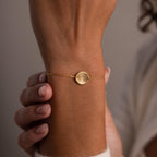 The Dainty Organic Fingerprint Bracelet, featuring a round disc charm, rests elegantly on a person’s wrist as their hands gently touch this unique keepsake jewelry piece.