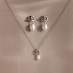The Pearl Knot Necklace & Earrings Set in Sterling Silver features a delicate knot design with dangling white pearls, perfect for wedding jewelry and elegantly displayed on a beige surface.