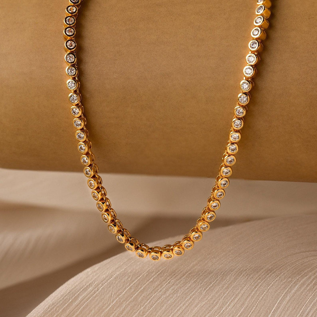 The Diamond Bezel Tennis Necklace & Bracelet Set features round bezel-set diamonds against a beige textured background, ideal for elegant pairings.
