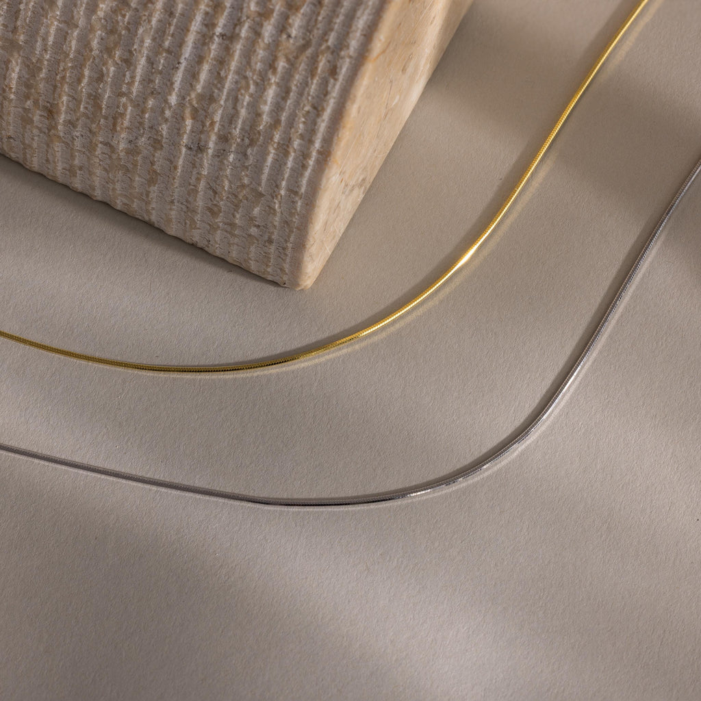 The Dainty Herringbone Necklace, in gold and silver, is displayed on a beige textured surface with a wooden block.