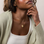 Woman wearing the Floral Handwriting Locket Necklace in 18K Gold, styled with a light sweater and white tank top, her hand resting on her chin.