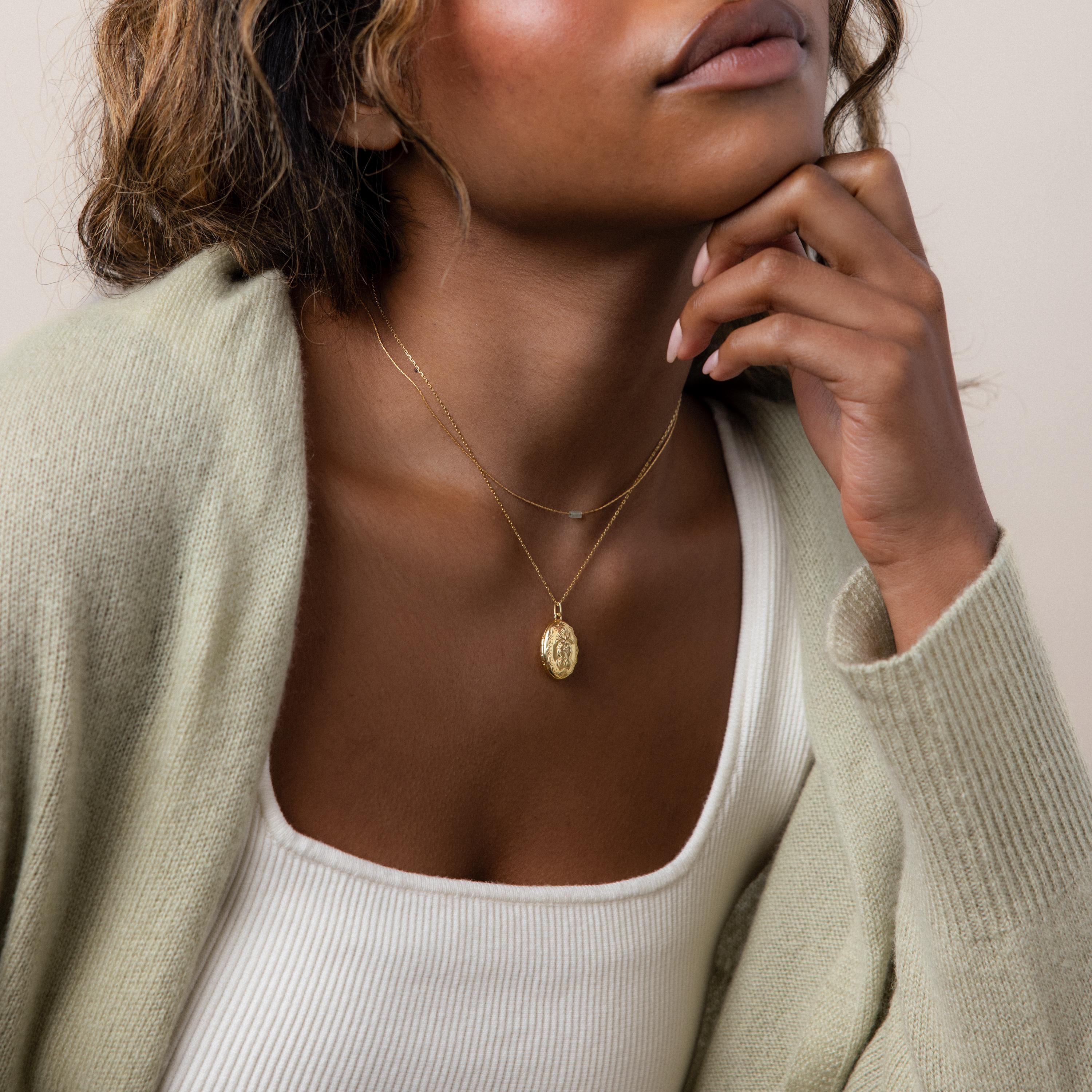 Woman wearing the Floral Handwriting Locket Necklace in 18K Gold, styled with a light sweater and white tank top, her hand resting on her chin.