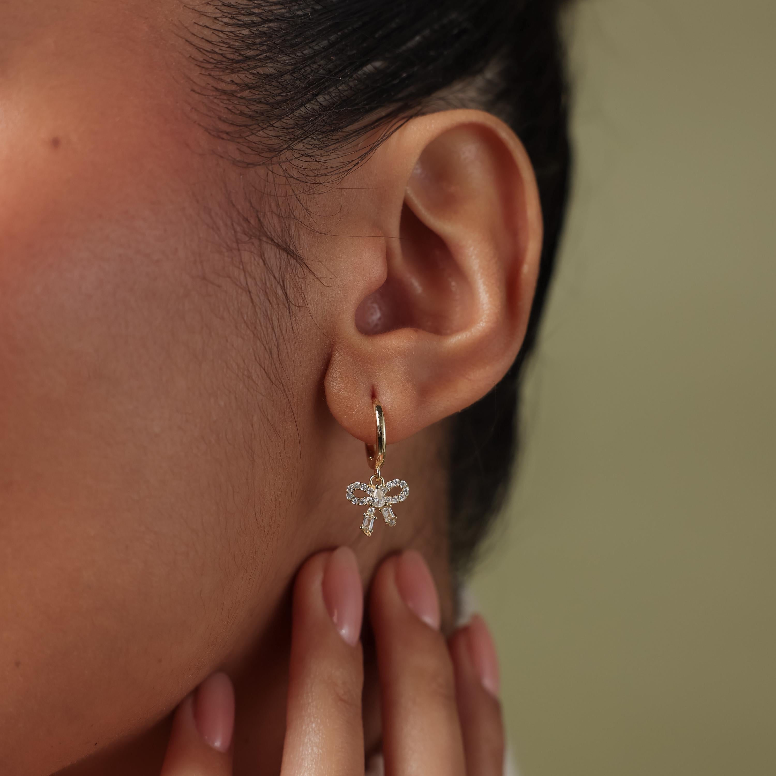 Close-up of an ear wearing a gold diamond bow hoop earrings against a neutral background.