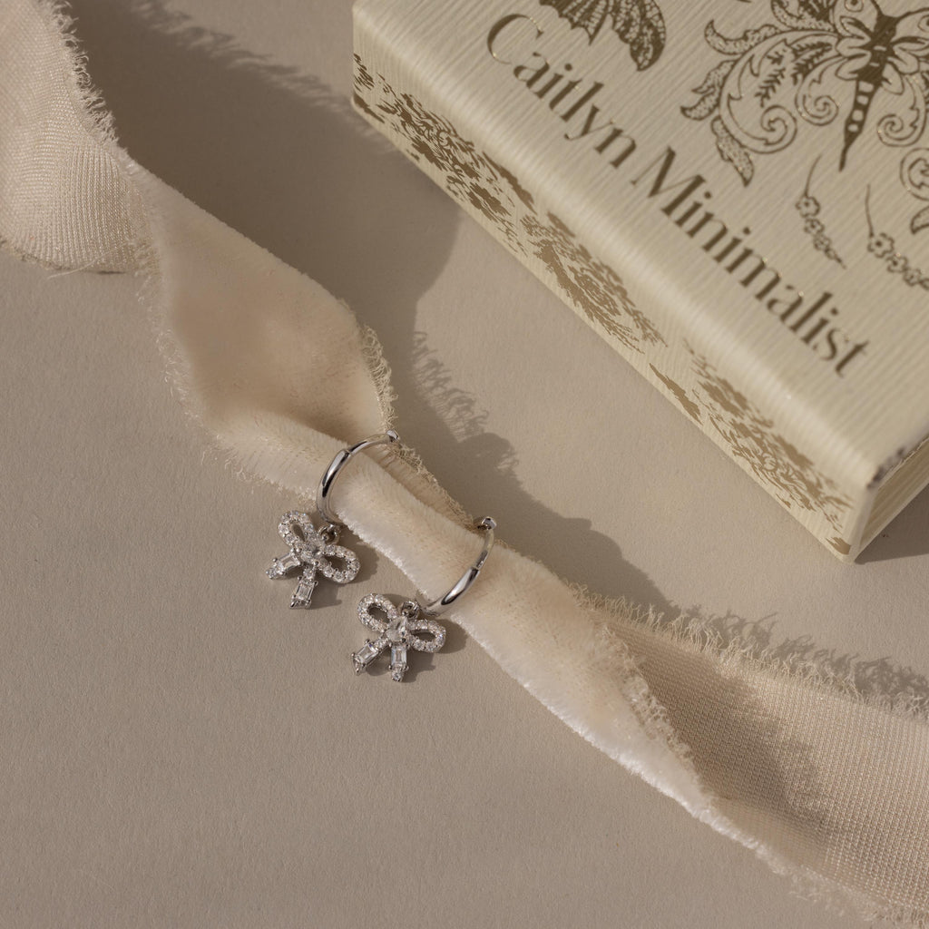 Flatlay of our Pave Bow Charm Hoops
 in Silver on a beige fabric with 'Caitlyn Minimalist' box in the background