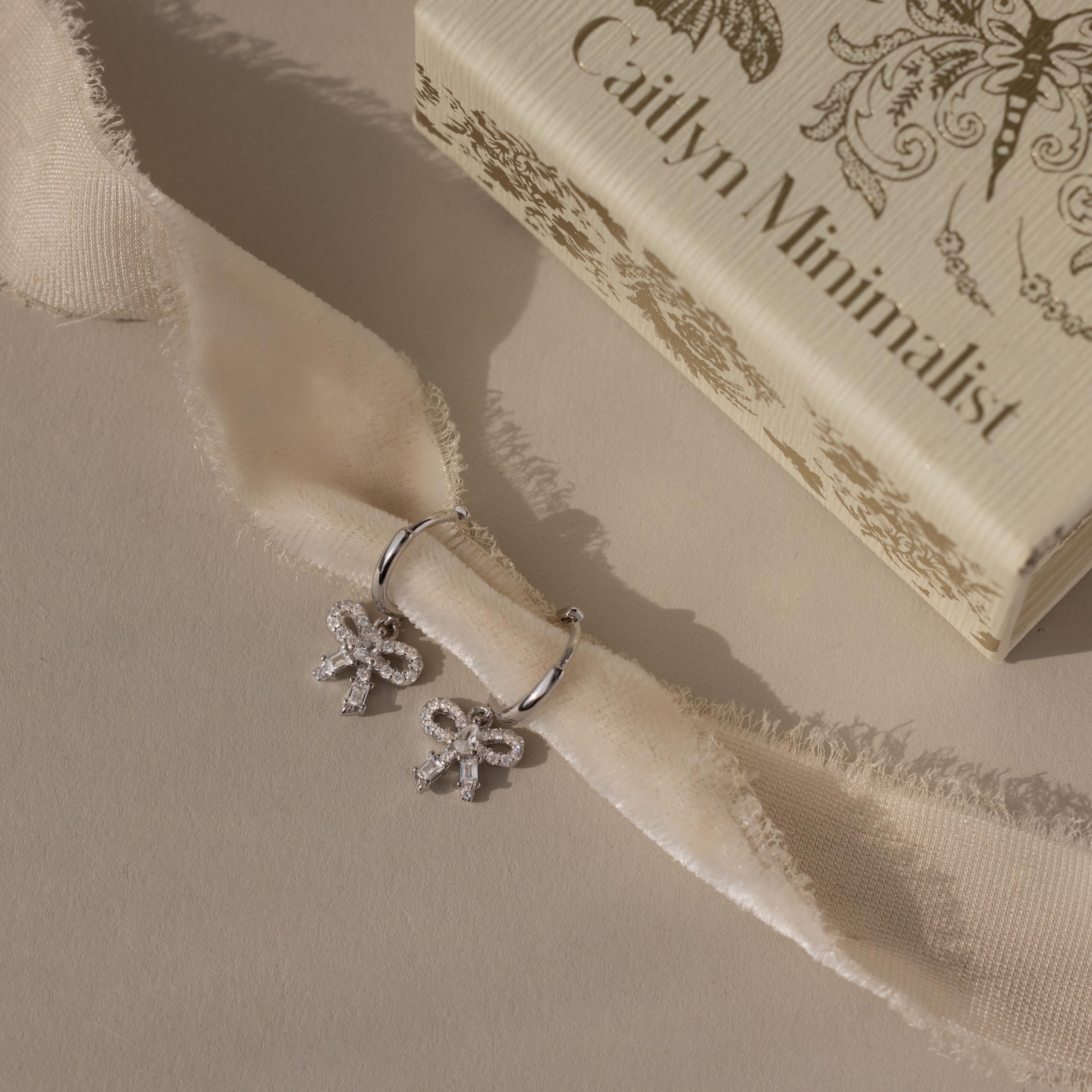 Flatlay of our Pave Bow Charm Hoops
 in Silver on a beige fabric with 'Caitlyn Minimalist' box in the background