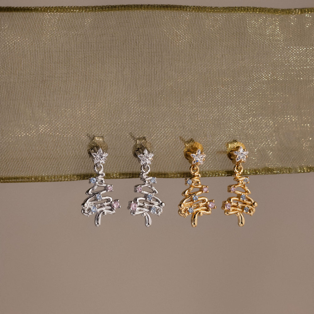 2 pairs of our Holiday Diamond Tree Studs in silver and gold on a beige fabric background
