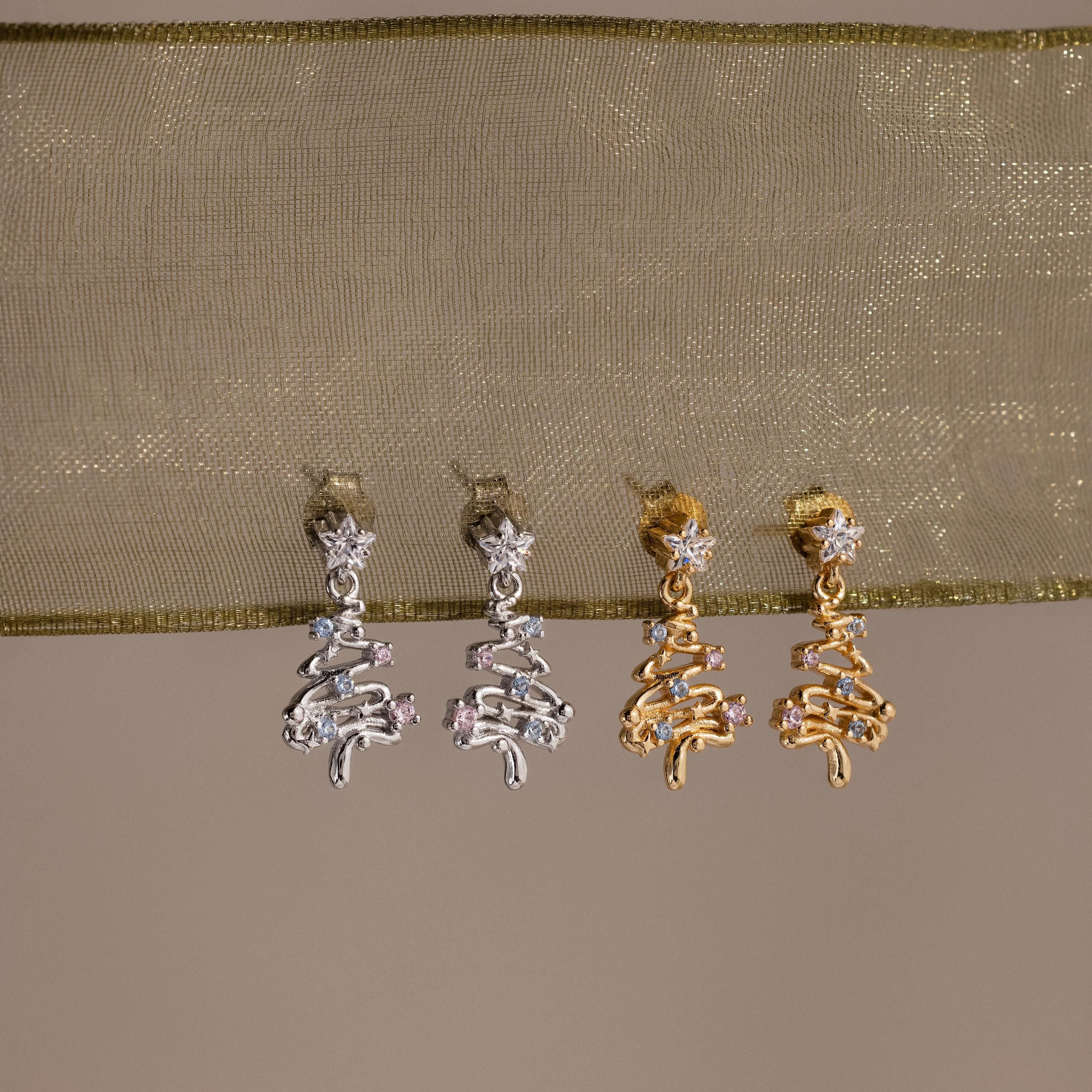 2 pairs of our Holiday Diamond Tree Studs in silver and gold on a beige fabric background