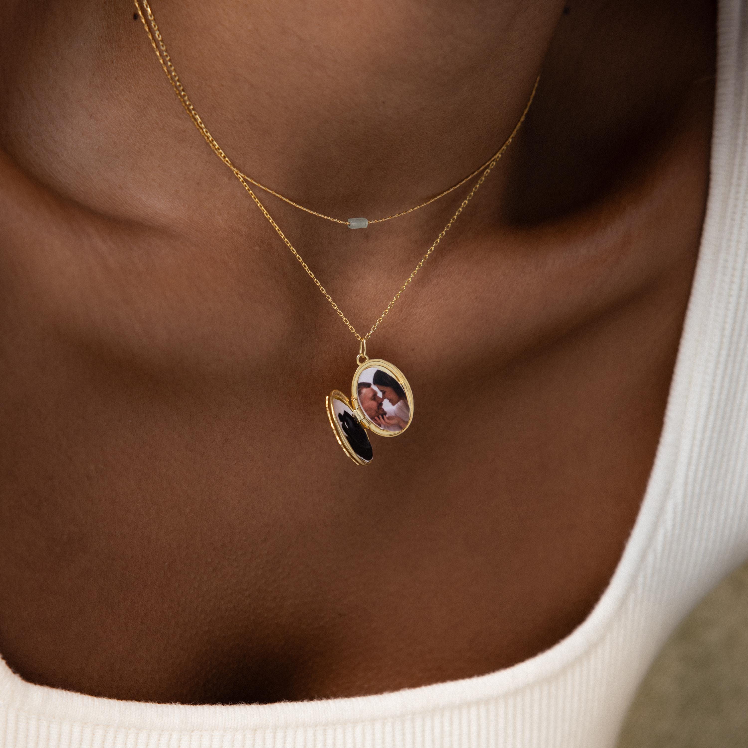 The Floral Handwriting Locket Necklace in 18K Gold is shown worn over a white top; a closeup highlights the delicate locket resting on the wearer’s neck and chest, with a photo tucked inside.