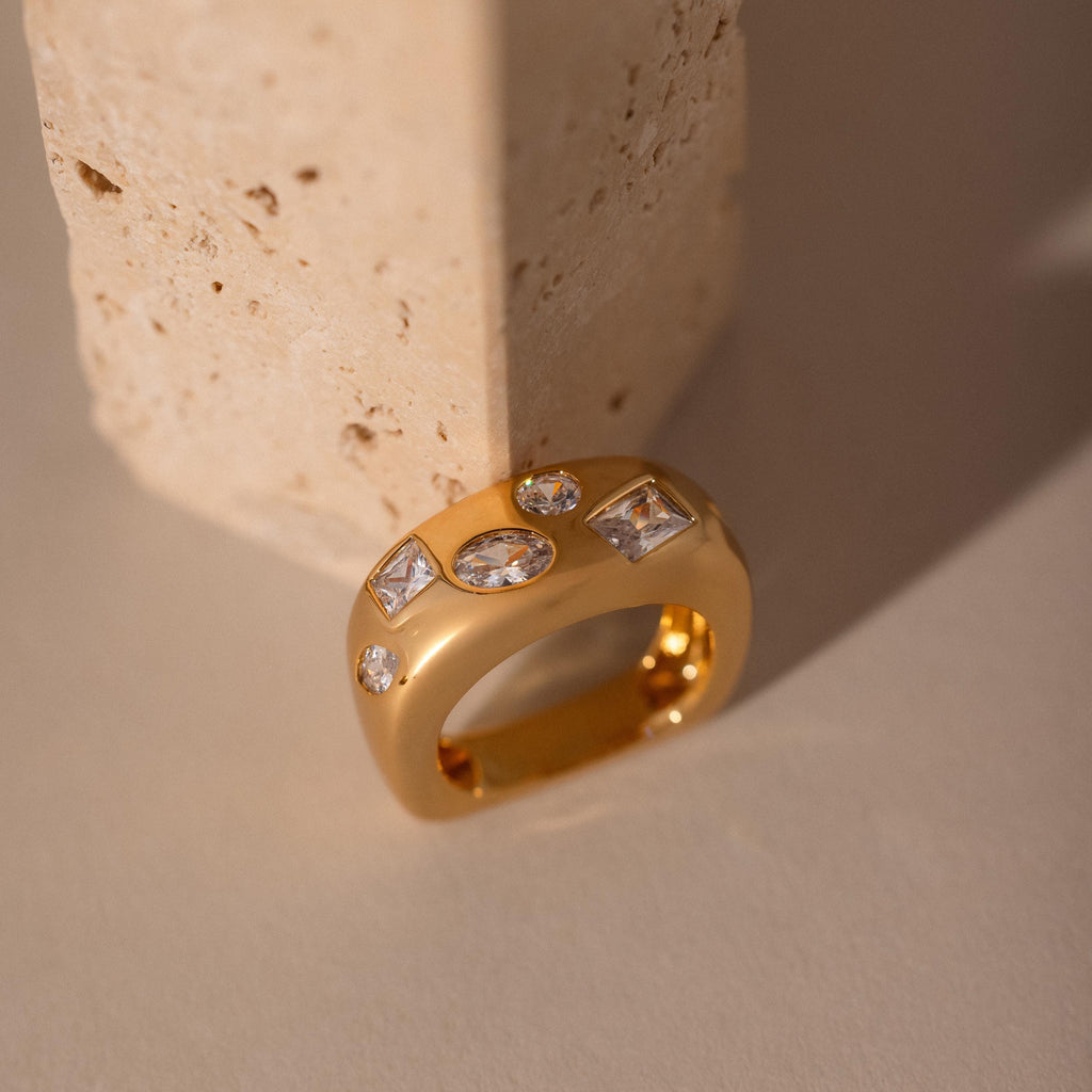 The Diamond Confetti Signet Ring features a chunky gold band with multiple clear gemstones, displayed against a textured beige stone block.