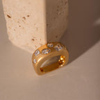 The Diamond Confetti Signet Ring features a chunky gold band with multiple clear gemstones, displayed against a textured beige stone block.