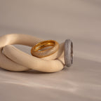 The Bold Pave Stacking Ring in silver rests alongside a gold wedding band on intertwined beige ceramic shapes against a soft backdrop.