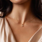 A woman wears the Rounded Pearl Flower Necklace, a delicate gold chain with a small floral pearl pendant, paired with a light beige satin top.