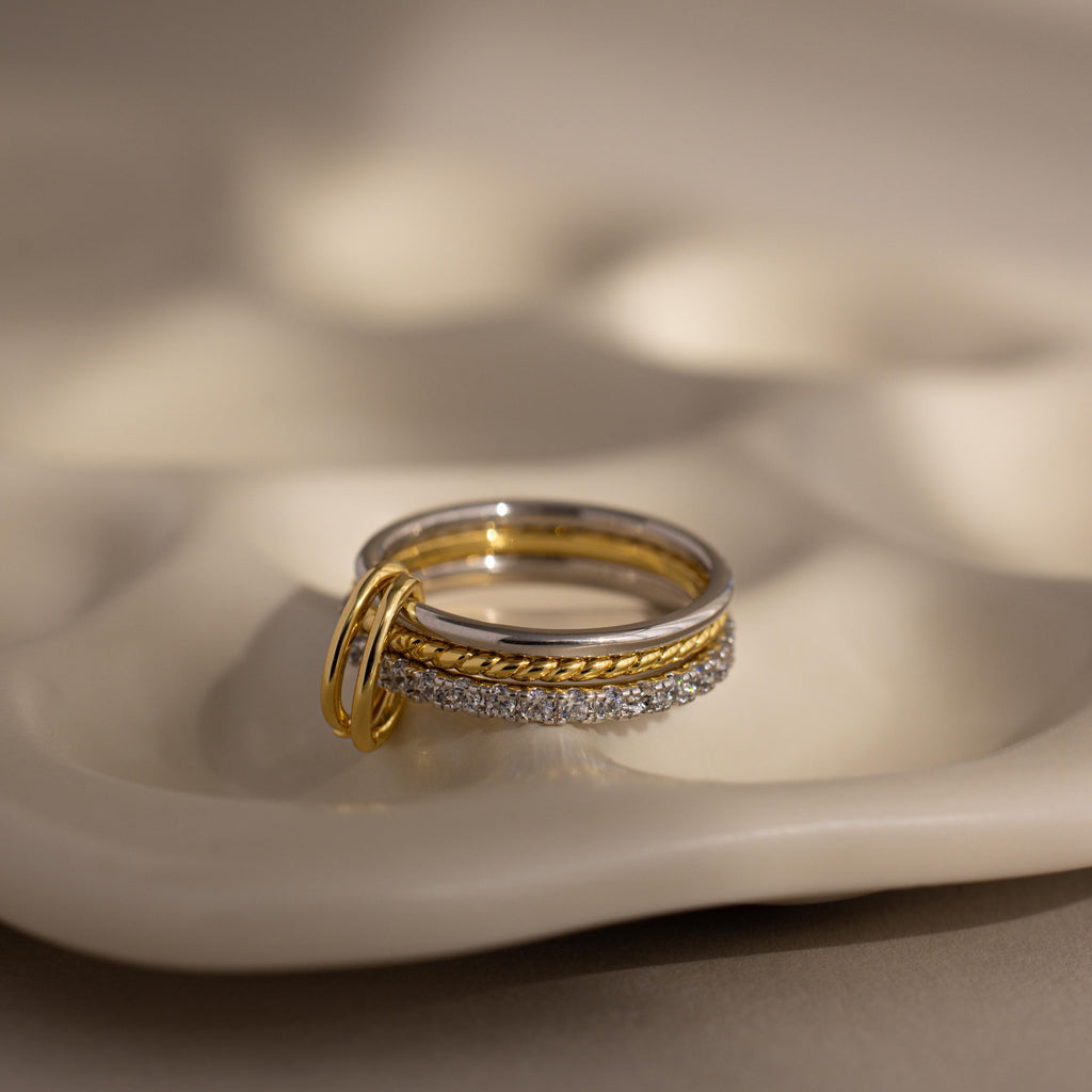 The Pave Mixed Metal Linked Ring Set features three stacked rings in gold and silver tones with sparkling pavé diamonds, elegantly displayed on a sculpted cream-colored dish—perfect for any occasion.