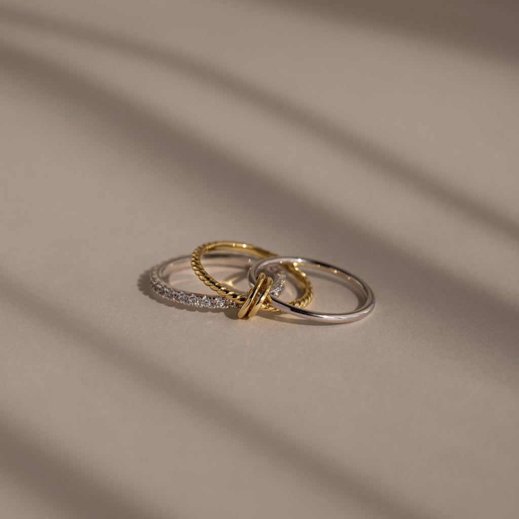 The Pave Mixed Metal Linked Ring Set, featuring gold, silver, and diamond-accented rings, rests on a smooth beige surface, highlighting its versatile jewelry style.