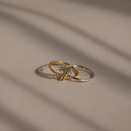 The Pave Mixed Metal Linked Ring Set, featuring gold, silver, and diamond-accented rings, rests on a smooth beige surface, highlighting its versatile jewelry style.