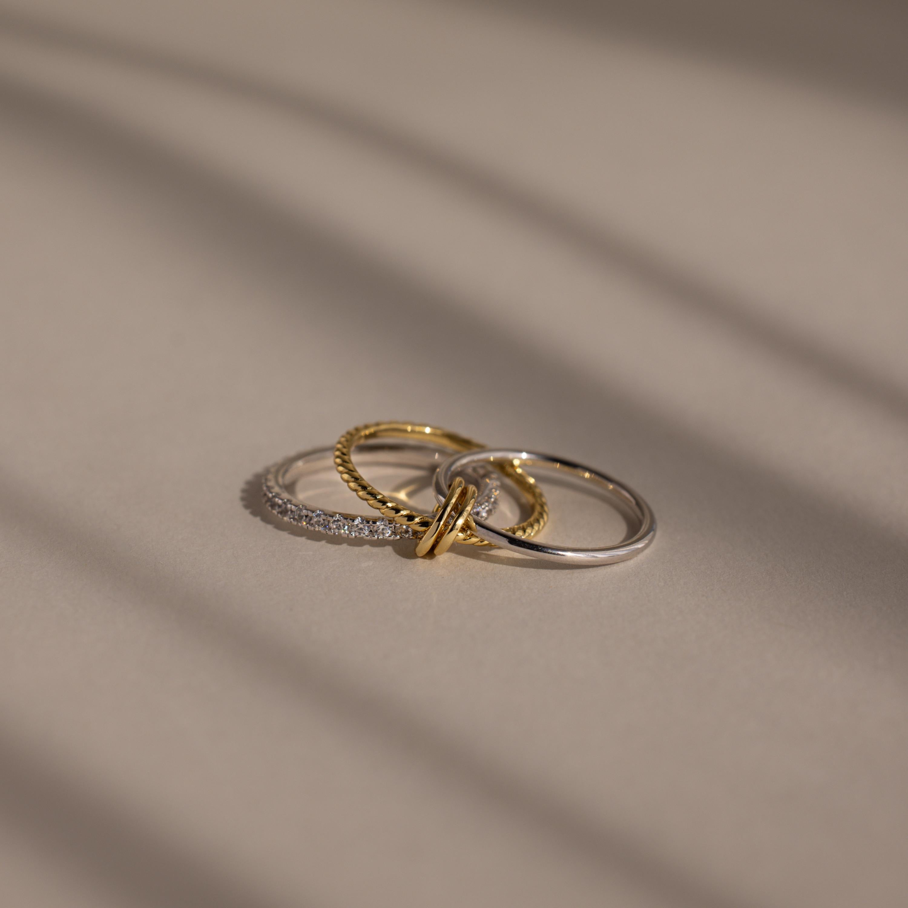The Pave Mixed Metal Linked Ring Set, featuring gold, silver, and diamond-accented rings, rests on a smooth beige surface, highlighting its versatile jewelry style.