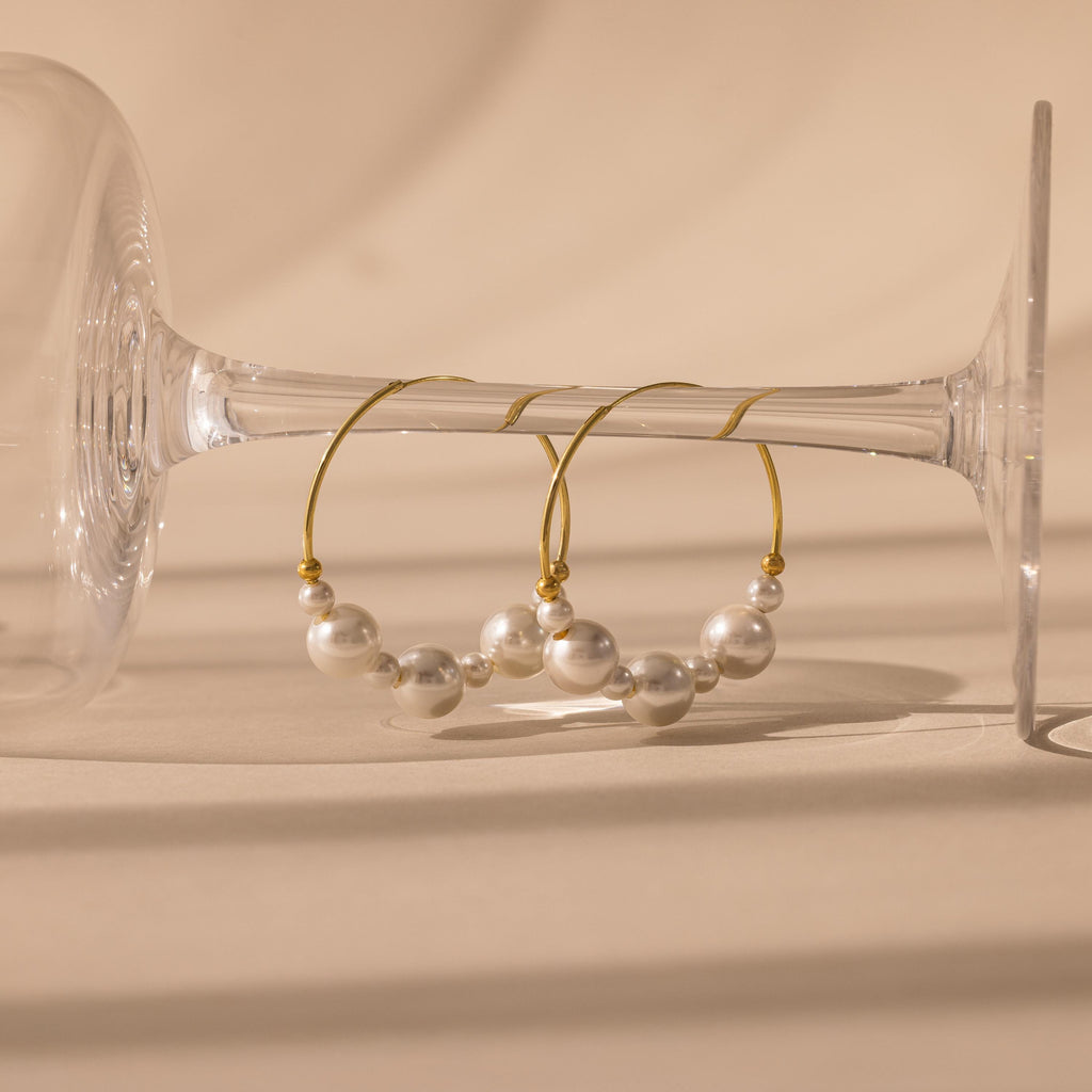 Statement Pearl Hoops in 18K Gold, adorned with white pearls, are displayed on the bowl of a clear, upside-down wine glass—an elegant showcase of quiet luxury.