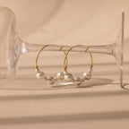 Statement Pearl Hoops in 18K Gold, adorned with white pearls, are displayed on the bowl of a clear, upside-down wine glass—an elegant showcase of quiet luxury.