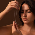 A woman holds up and admires the elegant Midnight Pearl Necklace in Sterling Silver, featuring a dark pendant, set against a brown background.