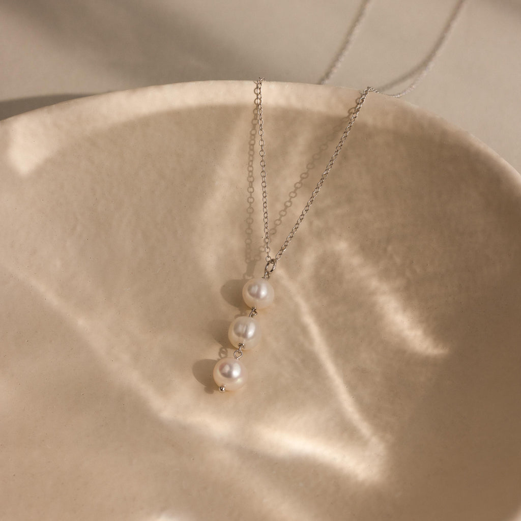 The Pearl Cascade Necklace in Sterling Silver, featuring three luminous pearls, rests on a textured, beige ceramic dish in soft light.