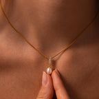 A hand holds the Diamond Pearl Droplet Necklace in 18K Gold, featuring a pearl and diamond pendant, against a woman's neck.