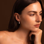 A woman with long brown hair wears elegant Diamond Pearl Droplet Earrings, gazing to the side with her chin resting on her hand.
