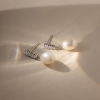 The Diamond Pearl Droplet Earrings in Sterling Silver rest on a cream surface, illuminated by soft natural light.