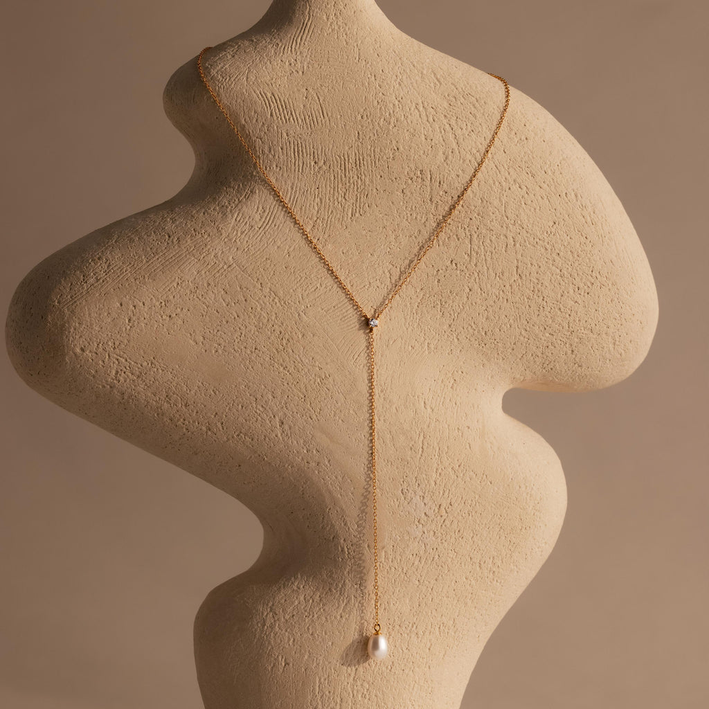 The Pearl Drop Lariat Necklace features a delicate gold chain and a pearl pendant, elegantly draped on a textured beige sculpture against a neutral background—perfect for those who love refined, elegant jewelry.
