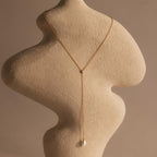 The Pearl Drop Lariat Necklace features a delicate gold chain and a pearl pendant, elegantly draped on a textured beige sculpture against a neutral background—perfect for those who love refined, elegant jewelry.