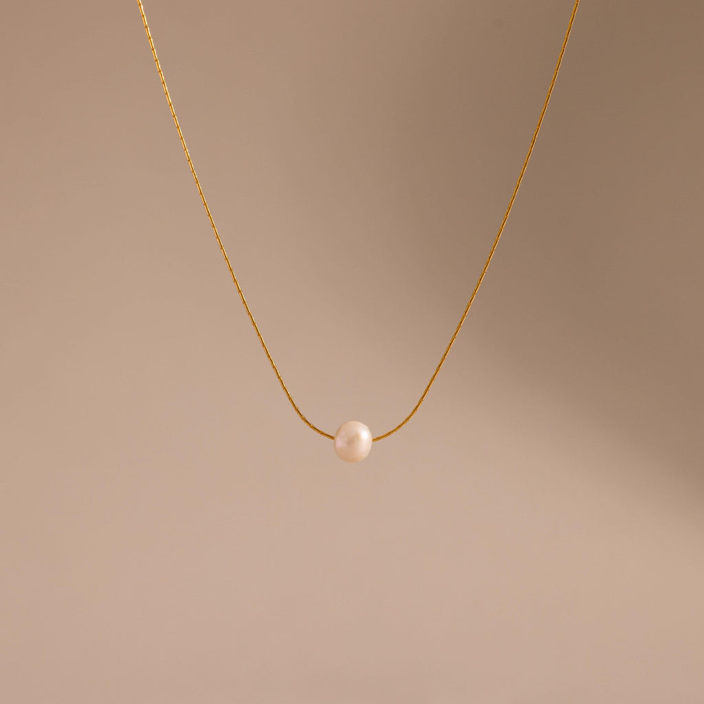 The Tiny Pearl Dot Necklace features a single white pearl pendant on a delicate gold chain, elegantly showcased against a soft beige background.
