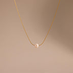 The Tiny Pearl Dot Necklace features a single white pearl pendant on a delicate gold chain, elegantly showcased against a soft beige background.