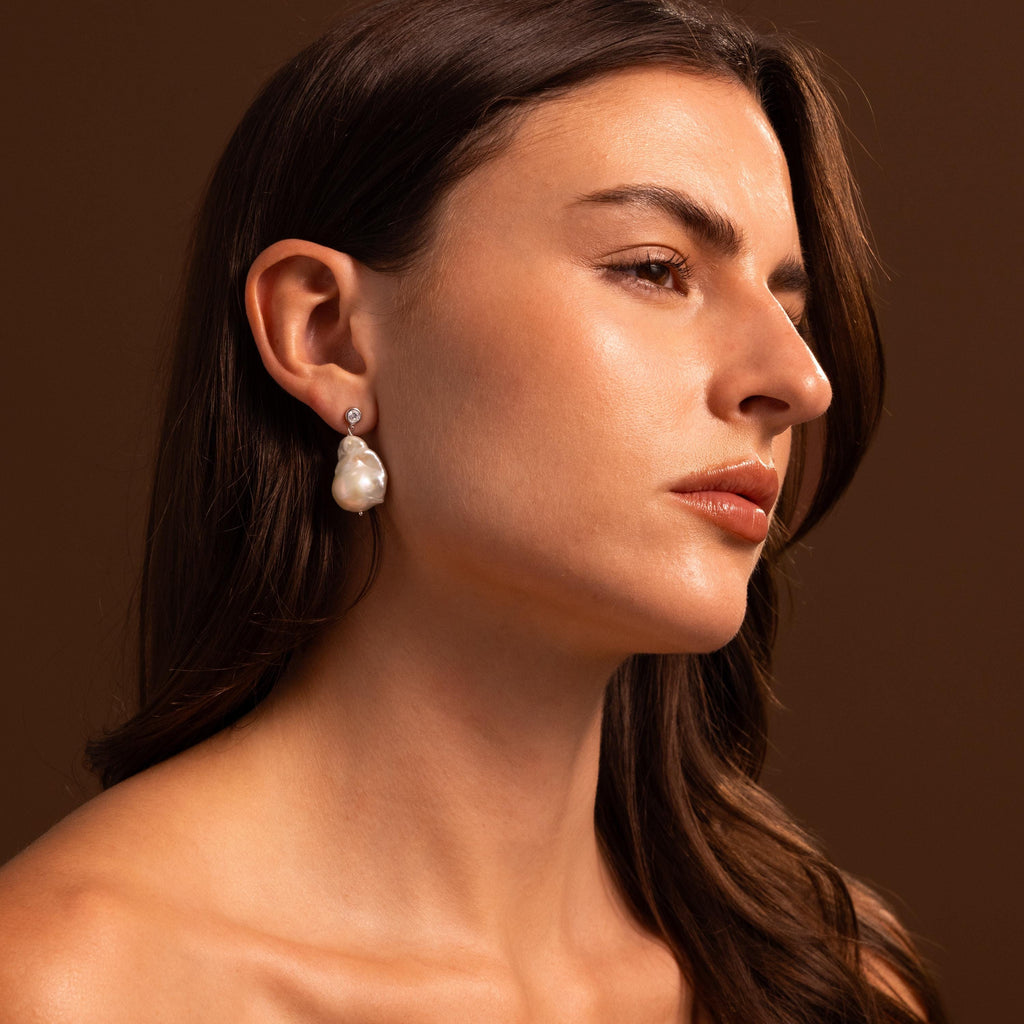 Woman with long brown hair gazes to the side, wearing Diamond Baroque Pearl Studs in Sterling Silver against a warm brown background.