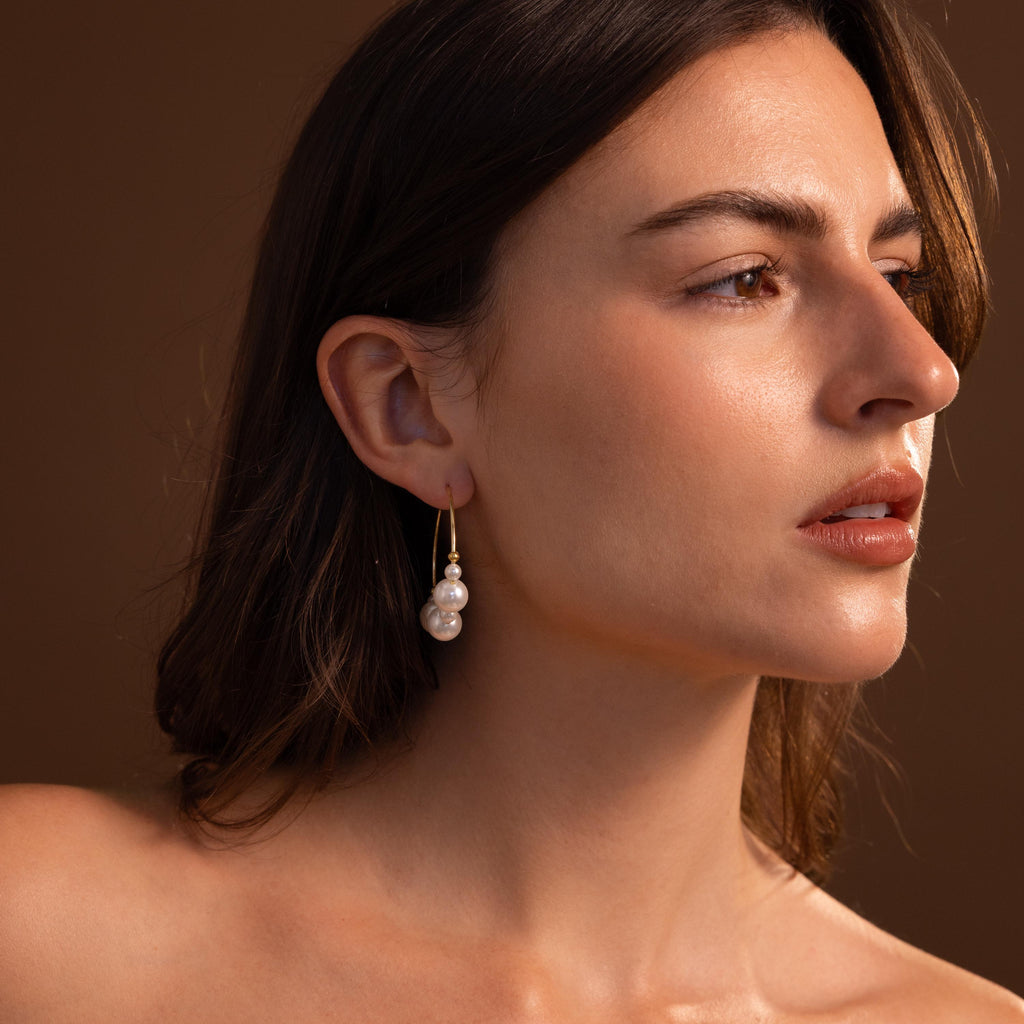 Woman with long brown hair wears Statement Pearl Hoops in 18K Gold, glancing to the side against a brown background.