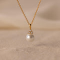 The Diamond Halo Pearl Necklace features a pearl pendant surrounded by small diamonds, elegantly showcased on a sandy surface—ideal for those who value timeless jewelry.
