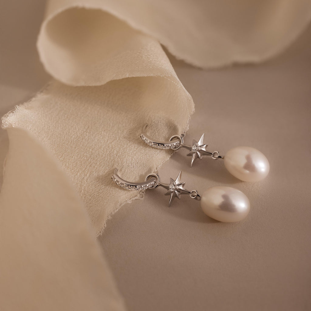 North Star Pearl Hoops adorned with star accents and delicate pearls, showcased on a soft beige fabric background.