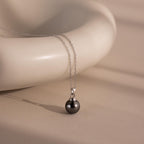 The Midnight Pearl Necklace in Sterling Silver, with its round dark pendant, is elegantly draped over a smooth cream ceramic surface—a stunning piece of jewelry.