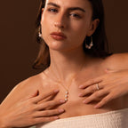 A woman poses with hands on chest against a brown background, showcasing the Pearl Cascade Necklace—paired with luminous pearl earrings and a matching ring.