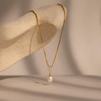 The Diamond Pearl Droplet Necklace in 18K Gold is elegantly draped over a textured beige sculpture, showcasing its gold chain and pearl pendant.