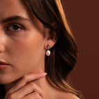 A woman with wavy brown hair, wearing the Midnight Pearl Drop Earrings in Sterling Silver—featuring elegant black accents—touches her chin against a brown background.