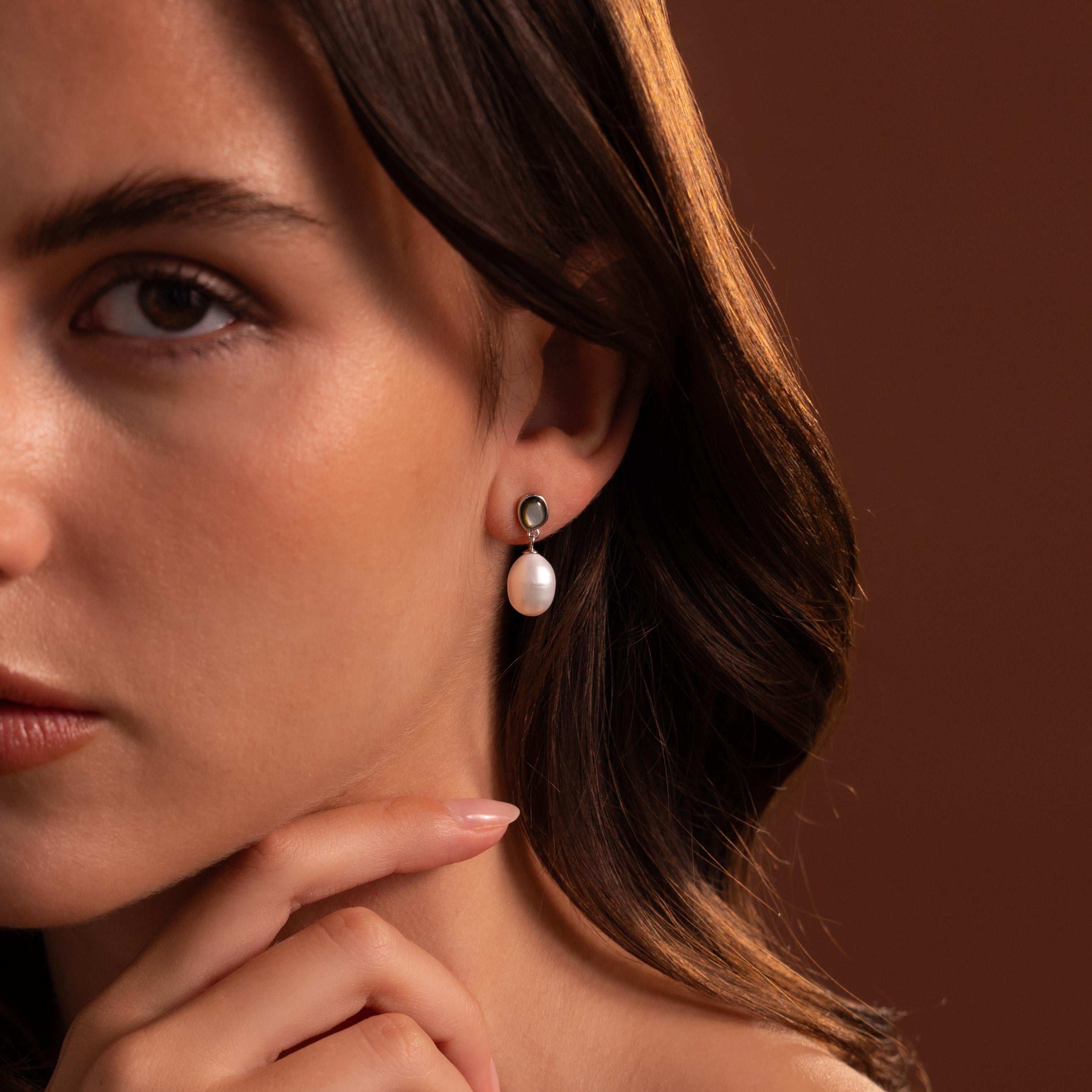 A woman with wavy brown hair, wearing the Midnight Pearl Drop Earrings in Sterling Silver—featuring elegant black accents—touches her chin against a brown background.