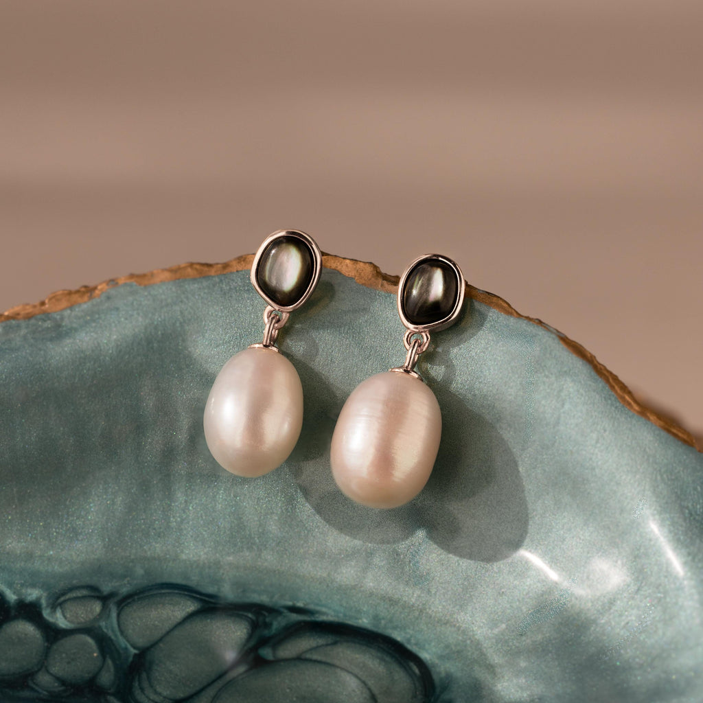 The Midnight Pearl Drop Earrings in Sterling Silver feature black oval studs with elegant white pearls and black accents, creating timeless jewelry. Shown on a blue ceramic surface.