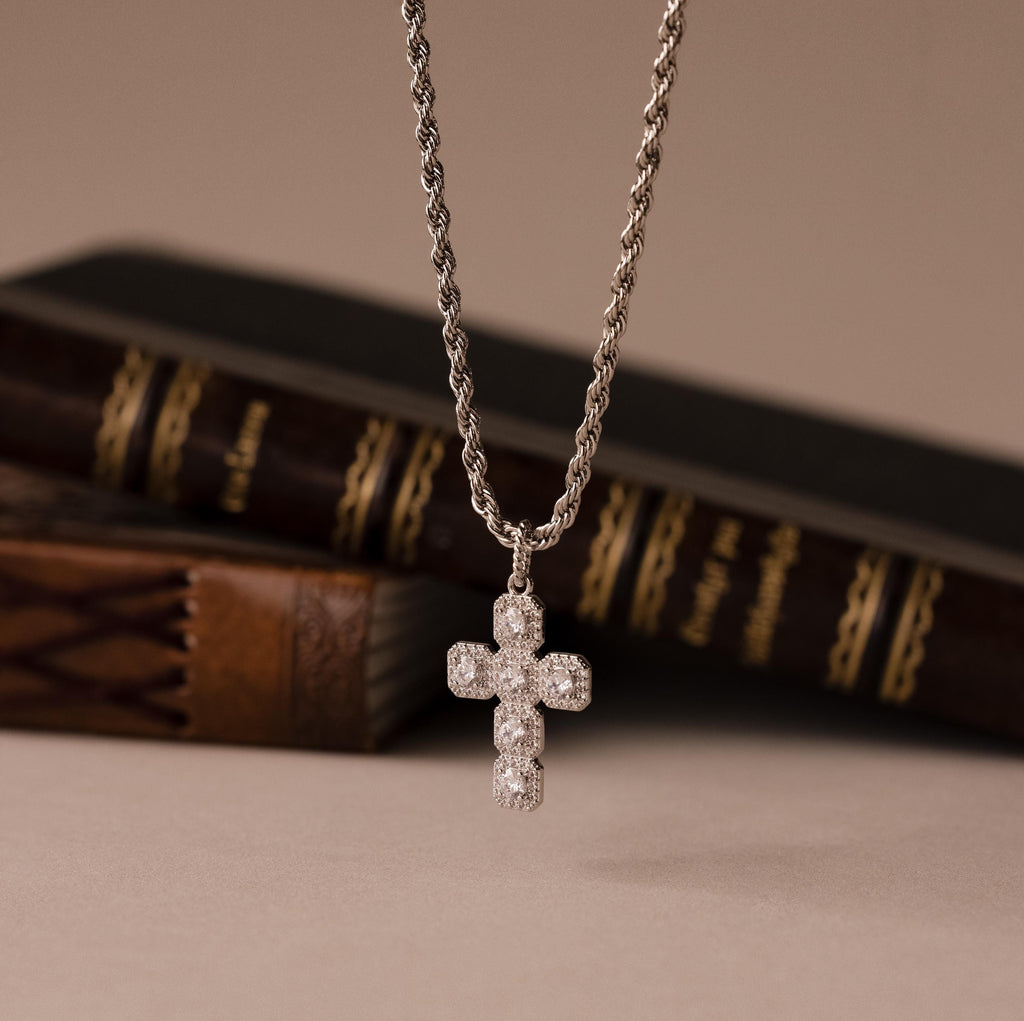The Majesty Cross Necklace in Silver features a sparkling stone-adorned cross pendant on a silver chain, elegantly displayed before stacked books.