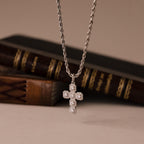 The Majesty Cross Necklace in Silver features a sparkling stone-adorned cross pendant on a silver chain, elegantly displayed before stacked books.