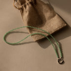 The Beaded Jade Clasp Necklace in Silver, ideal for layering, is displayed beside a brown burlap drawstring pouch on a beige surface.