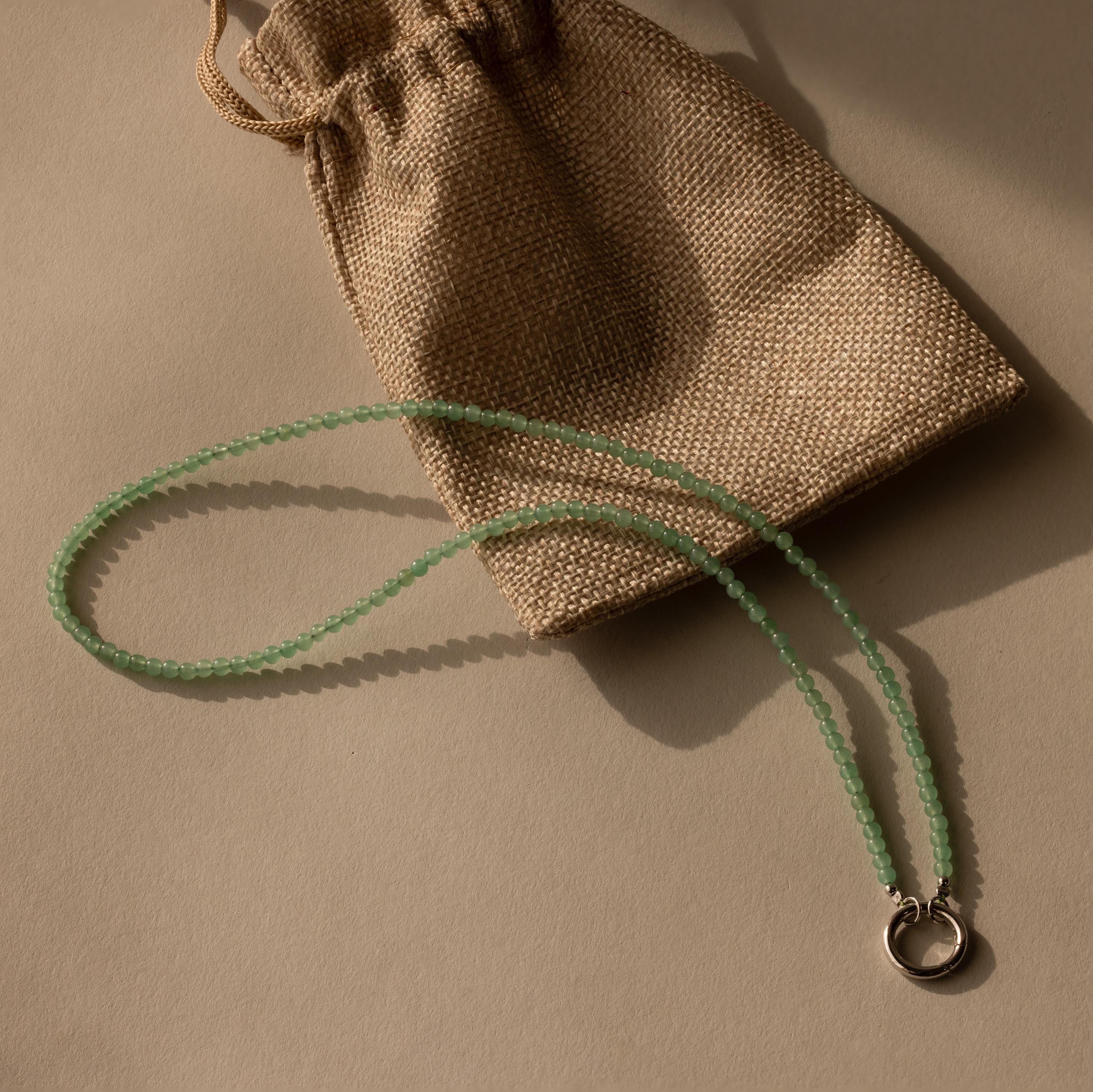 The Beaded Jade Clasp Necklace in Silver, ideal for layering, is displayed beside a brown burlap drawstring pouch on a beige surface.