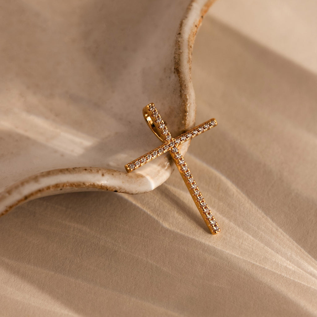 The Skinny Pave Cross Charm, featuring petite diamonds, rests on a textured beige ceramic surface and casts soft shadows—a timeless symbol of faith.
