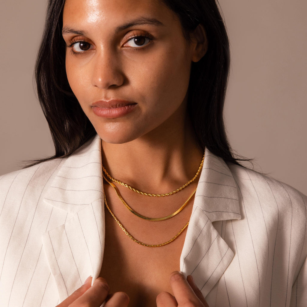 A woman in a white pinstripe blazer wears three layered gold chain necklaces, featuring the Bold Snake Chain Necklace in 18K Gold, showcasing modern jewelry while looking at the camera.
