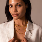 A woman in a white pinstripe blazer wears three layered gold chain necklaces, featuring the Bold Snake Chain Necklace in 18K Gold, showcasing modern jewelry while looking at the camera.