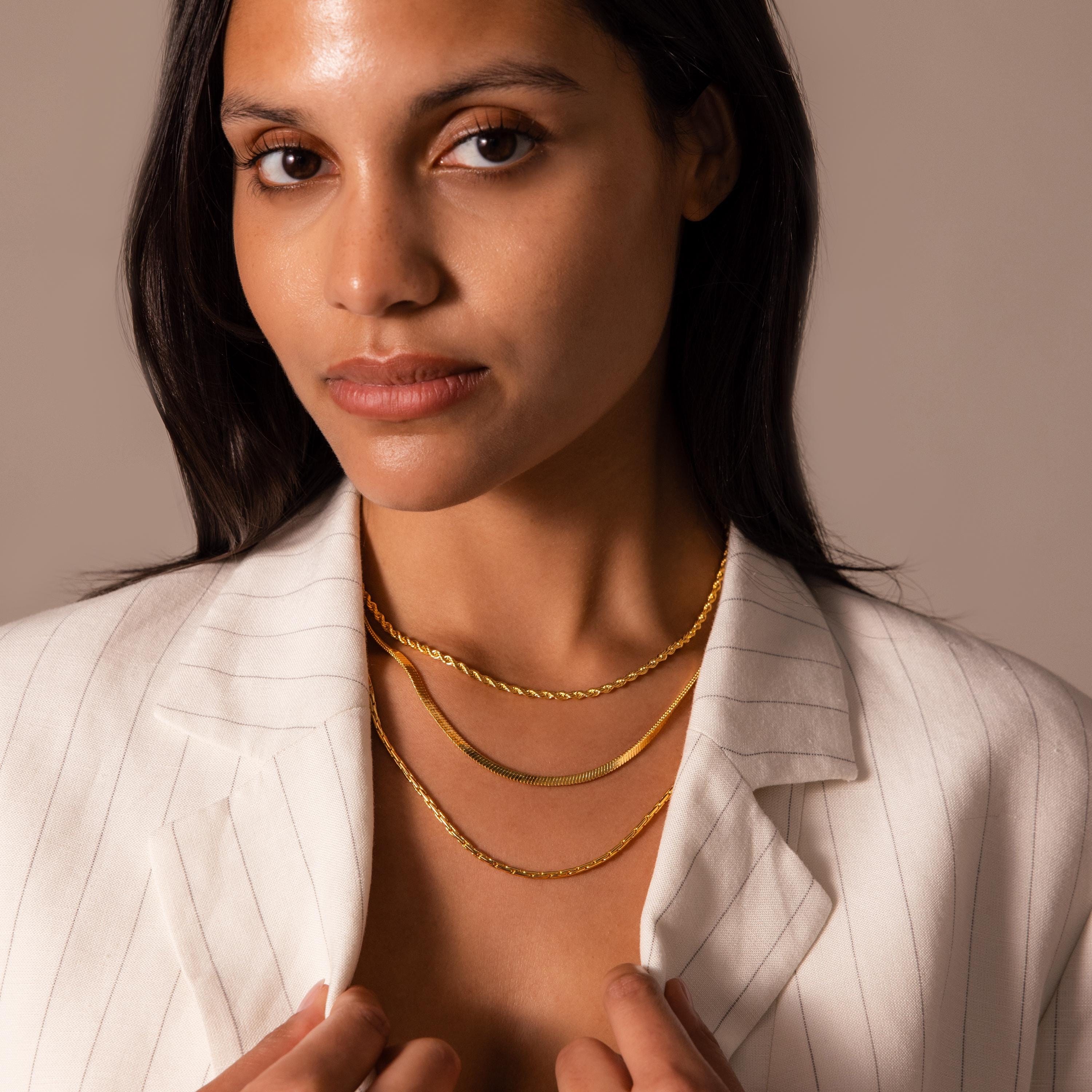 A woman in a white pinstripe blazer wears three layered gold chain necklaces, featuring the Bold Snake Chain Necklace in 18K Gold, showcasing modern jewelry while looking at the camera.